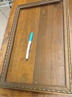 Full view of the wooden frame laid flat with a marker inside for scale, showing carved edge details and glass surface.