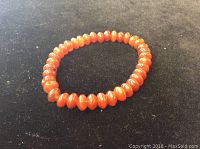 Close-up photo of an orange-red carnelian bead bracelet with smooth, oval-shaped beads on a stretchy cord against a black background.