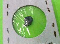 Rough alexandrite gemstone displayed under a transparent plastic film on a green circular area, showing its dark color and irregular shape.