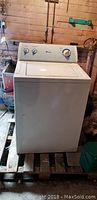 Full view of the white Amana top load washer placed on a wooden pallet in a basement or utility space.