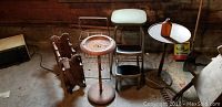 Photo of the vintage step stool, wire magazine rack beside it, metal pedestal ashtray with round tray top, and wooden accent table with white round top and a brown base in a dimly lit space.