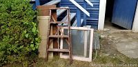 Wide view showing two vintage wood-framed windows, a wooden 4-foot step ladder, two wooden cabinet doors leaning against a blue siding building.