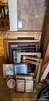 Full view of all picture frames stacked showing varying sizes and styles including large wooden, rectangular, and round frames.