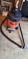 Full view of orange and black 10 gallon Shop Vac with attached hose and floor vacuum head on carpet.