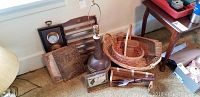 Photo showing multiple wicker baskets stacked, ceramic lamp, embossed tin, barometer, wooden spice rack, carved wooden panel, and wooden box. Items arranged on floor against wall and furniture.
