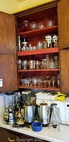 Four shelves filled with assorted clear glassware including stemware, jars, and novelty skeleton wine glasses. Countertop holds Osterizer blender, metal coffee percolators, Philips electric knife, blue bowl and other kitchenware. Visible wooden cabinet framing the shelves.