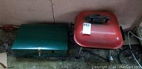 Photo of green Coleman portable camping stove in closed metal case beside red portable charcoal barbecue with lid and legs.