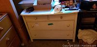 Full view of vintage wooden dresser painted light cream with 3 drawers and various items on top.