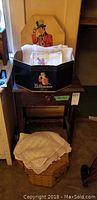 Photo showing vintage linens folded inside a black Baltimore box placed on top of a wooden table, with a vintage cardboard sign behind and a hat box with crocheted linens below.