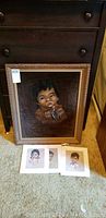 Framed black velvet portrait of Indigenous child leaning against a dark wooden cabinet with three small signed prints placed in front on a beige carpet.