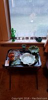 Full lot view showing assorted glass vases, decanter, ceramic serving dishes on vintage metal tray with folding stand by window
