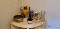 Overview photo showing nine assorted vases and a decorative metal bowl arranged on a corner shelf, various sizes and materials.