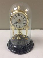Front view of gold-tone anniversary clock with white dial, black Roman numerals, pendulum, under a glass dome and marble base.