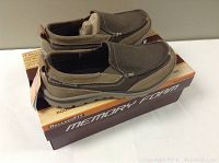 Pair of men's Skechers slip-on shoes in brown tones resting on labeled shoe box showing 'Relaxed Fit' and 'Memory Foam' features.