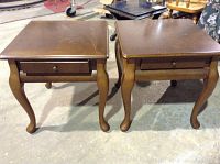 Photo showing two matching wooden end tables with single drawers and curved legs, placed side by side on a concrete floor. Visible surface scratches on tabletops.