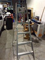 Photo showing an aluminum domestic step ladder standing in a storage area with multiple steps, support braces, no visible damage.