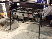 Front view of Camp Chef Xpedition 3X 3-burner stove showing steel frame, cooking grid, control knobs, and propane hose