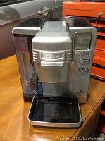 Silver Cuisinart Keurig coffee maker viewed from front and slightly left, showing water reservoir, digital interface, buttons, and drip tray.