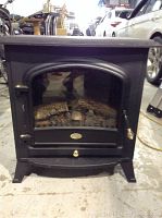 Front view of a black stove-style Dimplex electric heater with glass door and brass handle, showing faux logs inside. Positioned on a concrete floor inside a warehouse or garage with cars and furniture in background.