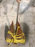 Photo showing a rusty metal boat anchor with two triangular flukes tied together by a rod shank, with yellow rope wrapped around for securing.
