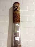 Rolled area rug standing vertically against a wall, wrapped partially in transparent protective plastic, showing detailed ornamental pattern in red, beige, gold, and brown.