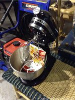 Black KitchenAid stand mixer with attachments including stainless steel bowl, wire whisk, and flat beater, placed on a wicker table in a workshop setting.