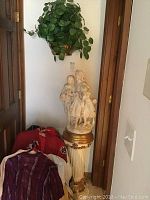 Resin statue of three girls on white and gold pedestal with faux green wall plant above, displayed in corner near doorways.