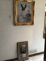 Two framed pictures displayed on a wall, one mounted higher and one leaning below it near a vent. The larger top picture shows the eagle and nest, the lower picture shows the church and cross.