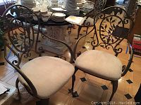 Two metal dining chairs with intricate leaf-patterned backs and white cushion seats photographed on hardwood tiled floor with various household items in the background.