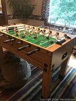 Side perspective showing the full wooden foosball table with player figures on rods and scoreboard at the back, set on a patterned rug indoors near window.