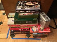 Photo of stacked board games including Mastermind, Rummikub, Memory, Charade, Bibleopoly, Dawg-opoly, Irishopoly, Whatzit?, Lord of the Rings Risk, and others; plus metal box of dominos on right side