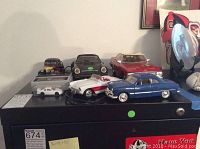 Photo showing seven die cast cars including a red Mercedes and several classic car models, along with one remote control car and its wired controller on a black surface.
