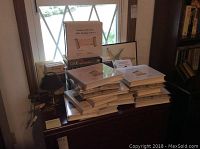 Photo of multiple white binders stacked on a wooden cabinet near a window with a cardboard box and framed artwork behind