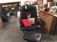 Black leather chair with Georgia Bulldog logo, displayed with foot stool, pillow, and packaged furniture protector in a room with furniture and boxed items.