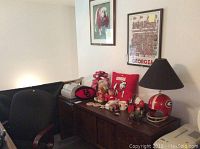 View of two framed Georgia Bulldogs pictures on wall above a wooden cabinet with multiple Bulldogs memorabilia including lamp with helmet base, stuffed animals, football, pillow, and small collectibles.