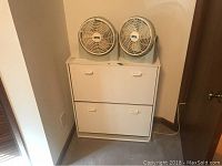 White wooden shoe cabinet with two compartments and two Lasko fans sitting on top.