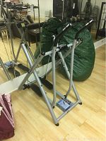 Full view of the Tony Littles Gazelle Freestyle stepper machine in silver with black pedals and foam handles in a room with mirrored wall and wood floor.