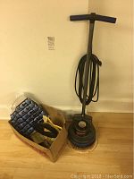 Full view of Oreck Orbiter XL floor buffer cleaner with cord wrapped around handle and box of attachments including cleaning pads and brushes on a hardwood floor.