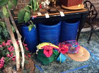 Overall view showing two large blue foam pool noodles under a black bench with a green inflatable pool float with red/yellow claws and a pink mesh bag in front, along with natural woven sunhat on the floor.
