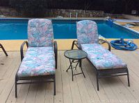 Photo of two pastel padded adjustable lounger chairs and small round side table by a pool on wooden decking.