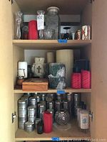 Cabinet shelves showing red and white candles of various sizes, glass and metal candle holders, a glass jar with lid, and incense in metal tin containers.