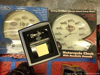 Two boxed wall clocks, one Ford with twelve classic Ford vehicles that sound motor running every hour and one Harley-Davidson that sounds motorcycle revving every hour, with a Harley-Davidson desk set box between them.