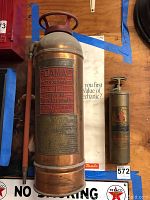 Two vintage fire extinguishers on a wooden surface; larger Foamac extinguisher with pressure gauge and hose, and smaller Fyr Fyter extinguisher with colorful label.