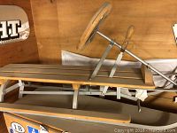 Wooden Swiss sled showing natural wood slatted seat and metal frame with steering wheel and wooden brake lever.