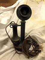 Front view of black metal candlestick phone showing mouthpiece, earpiece hook, and coiled fabric-covered cord with modular connector.