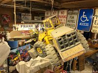 Front side view of dusty yellow Tonka Mighty Mighty loader toy truck hanging from rafters, visible vintage signs and model airplane in background