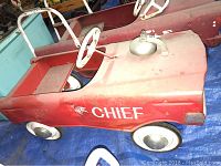Photo of smaller red fire department pedal car showing side view with white steering wheel, metal wheels with hubcaps, and 'CHIEF' marking.
