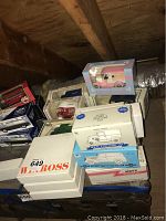 Stacked boxed collectible die-cast cars and banks in various white, blue, and red boxes on a wooden surface under a wooden beam.