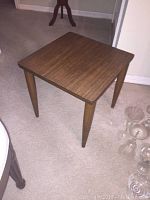 Photo of one wooden square side table showing tabletop and four tapered legs on carpeted floor