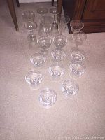Photo showing 18 pieces of clear assorted glassware including tall wine glasses, medium wine glasses, small wine glasses, and dessert cups placed on carpet.